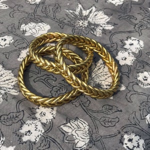 Puede incluir: Tres pulseras de brazalete trenzadas de color dorado apiladas una encima de la otra. Las pulseras est&aacute;n hechas de un material grueso con forma de cuerda.