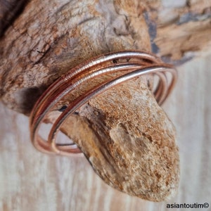May include: A set of copper-colored bangles, with a textured surface, are arranged on a piece of driftwood. The bangles are circular and appear to be made of metal. The wood has a natural, weathered appearance.