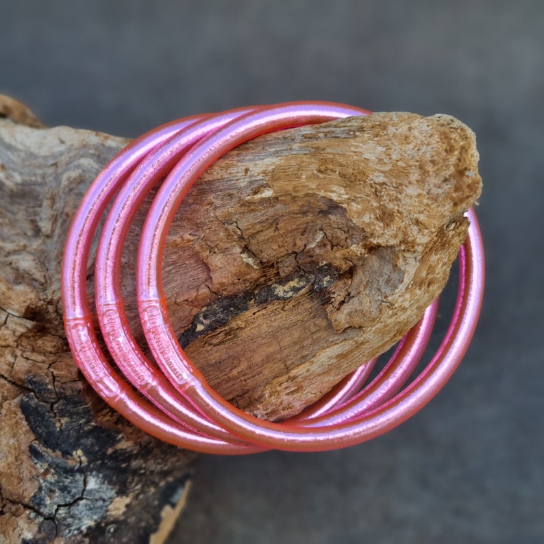 K&ouml;nnte beinhalten: Vier rosa, schillernde Armreifen sind auf einem St&uuml;ck Treibholz aufgewickelt. Die Armreifen haben ein glattes, abgerundetes Design und ein gl&auml;nzendes Finish. Das Holz ist hellbraun mit rauer Textur, und der Hintergrund ist ein ged&auml;mpftes Grau.