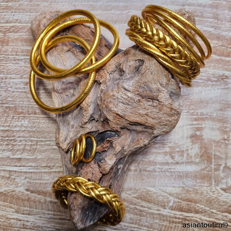 May include: A set of gold-colored leather bracelets, some with a braided design, on a piece of driftwood.