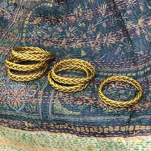 May include: Four gold braided rings stacked on top of each other on a patterned fabric background.