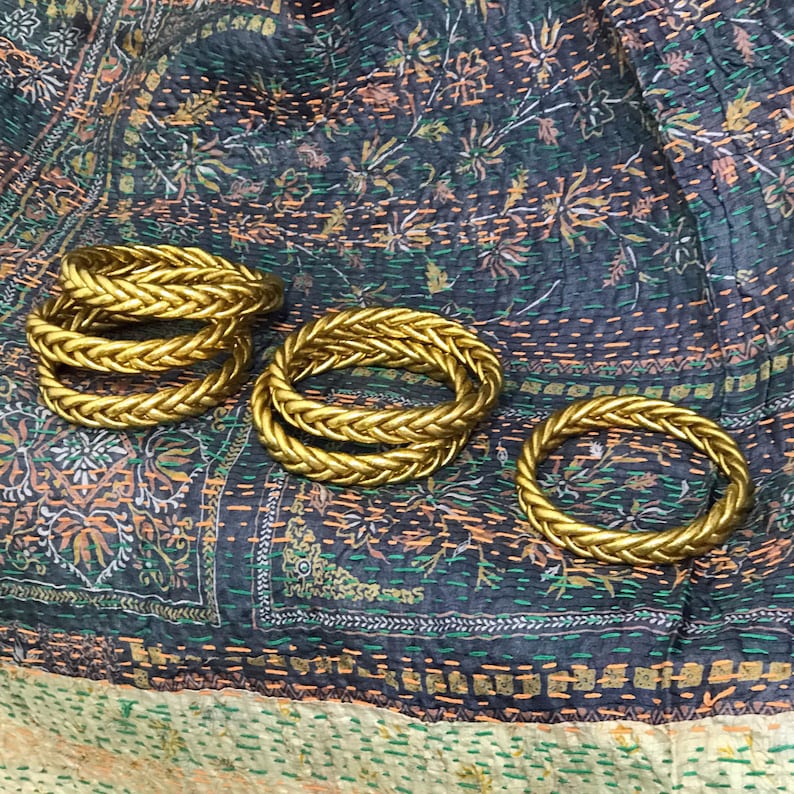 May include: Four gold braided rings stacked on top of each other on a patterned fabric background.