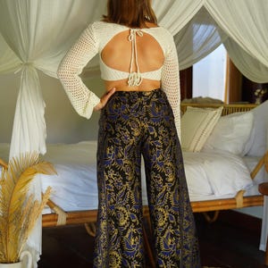 May include: A woman wearing a white crochet top with a keyhole back and black wide-leg pants with a blue and gold floral pattern.