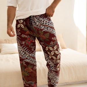 May include: Burgundy trousers with a detailed batik pattern featuring floral and cloud-like motifs. The trousers have a drawstring waist and are paired with a white t-shirt. The style is casual and comfortable.