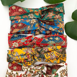 May include: Four colourful patterned headbands with a twisted knot design. The headbands are made of a silky fabric with a floral and fan-shaped pattern in shades of red, blue, yellow, and white.