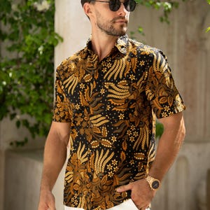 May include: Short-sleeved button-down shirt with a black background and a gold floral pattern. The shirt is paired with white pants. The shirt's design features stylized leaves and star-like shapes. The model is wearing sunglasses and a gold watch.
