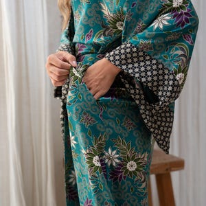 Op de afbeelding: Teal kimono-stijl badjas met een bloemenpatroon in wit, paars en groen. De badjas heeft wijde mouwen met een zwart-wit geometrisch ontwerp aan de manchetten. De persoon draagt de badjas en heeft de handen in de zakken.