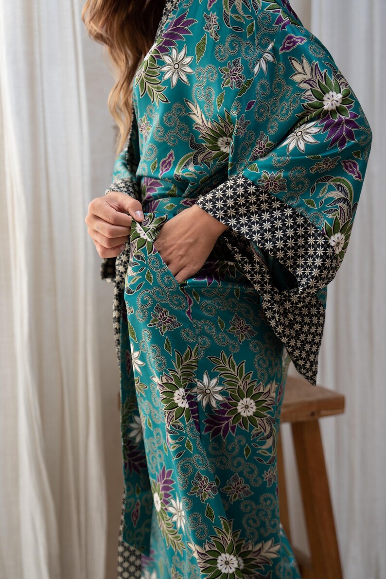 Op de afbeelding: Teal kimono-stijl badjas met een bloemenpatroon in wit, paars en groen. De badjas heeft wijde mouwen met een zwart-wit geometrisch ontwerp aan de manchetten. De persoon draagt de badjas en heeft de handen in de zakken.