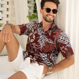 Può includere: Una camicia bordeaux a maniche corte con un motivo bianco e blu, abbinata a pantaloncini bianchi. La persona indossa occhiali da sole e un orologio d'oro. Il design della camicia presenta forme e spirali astratte.