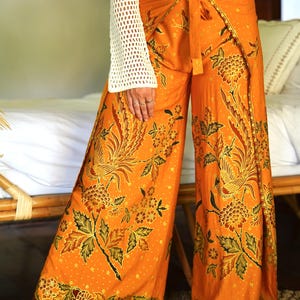 Op de afbeelding: Oranje wijde broek met een bloemenprint met vogels en bladeren. De broek heeft een wikkel-stijl met een strik in de taille.