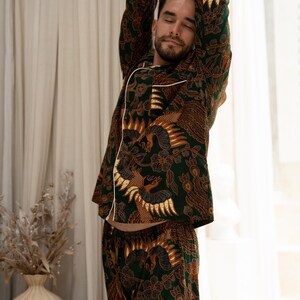 Puede incluir: Conjunto de pijama con estampado verde y marr&oacute;n con ribete blanco. El conjunto incluye una parte superior de manga larga y pantalones a juego. El patr&oacute;n presenta un dise&ntilde;o detallado con figuras de p&aacute;jaros y elementos florales. La persona de la imagen se estira.