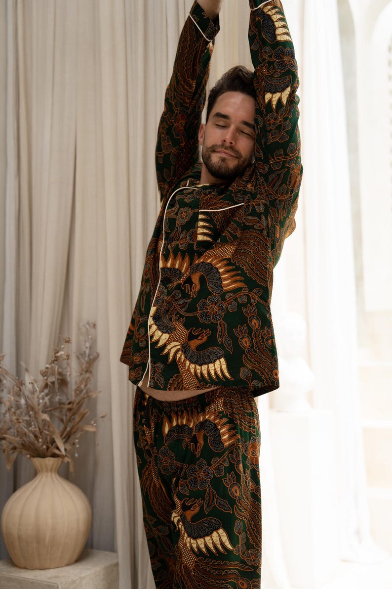 Puede incluir: Conjunto de pijama con estampado verde y marr&oacute;n con ribete blanco. El conjunto incluye una parte superior de manga larga y pantalones a juego. El patr&oacute;n presenta un dise&ntilde;o detallado con figuras de p&aacute;jaros y elementos florales. La persona de la imagen se estira.