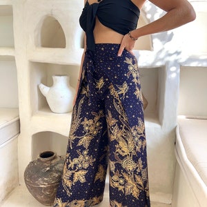 Puede incluir: Una mujer lleva un top corto negro y pantalones anchos con estampado floral dorado y azul. Los pantalones tienen un estilo de envoltura y están hechos de una tela suave y fluida.
