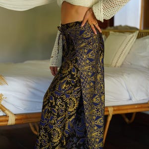 May include: A woman wearing a white crochet top and a long, blue and gold patterned wrap skirt. The skirt has a floral design and is tied at the waist.