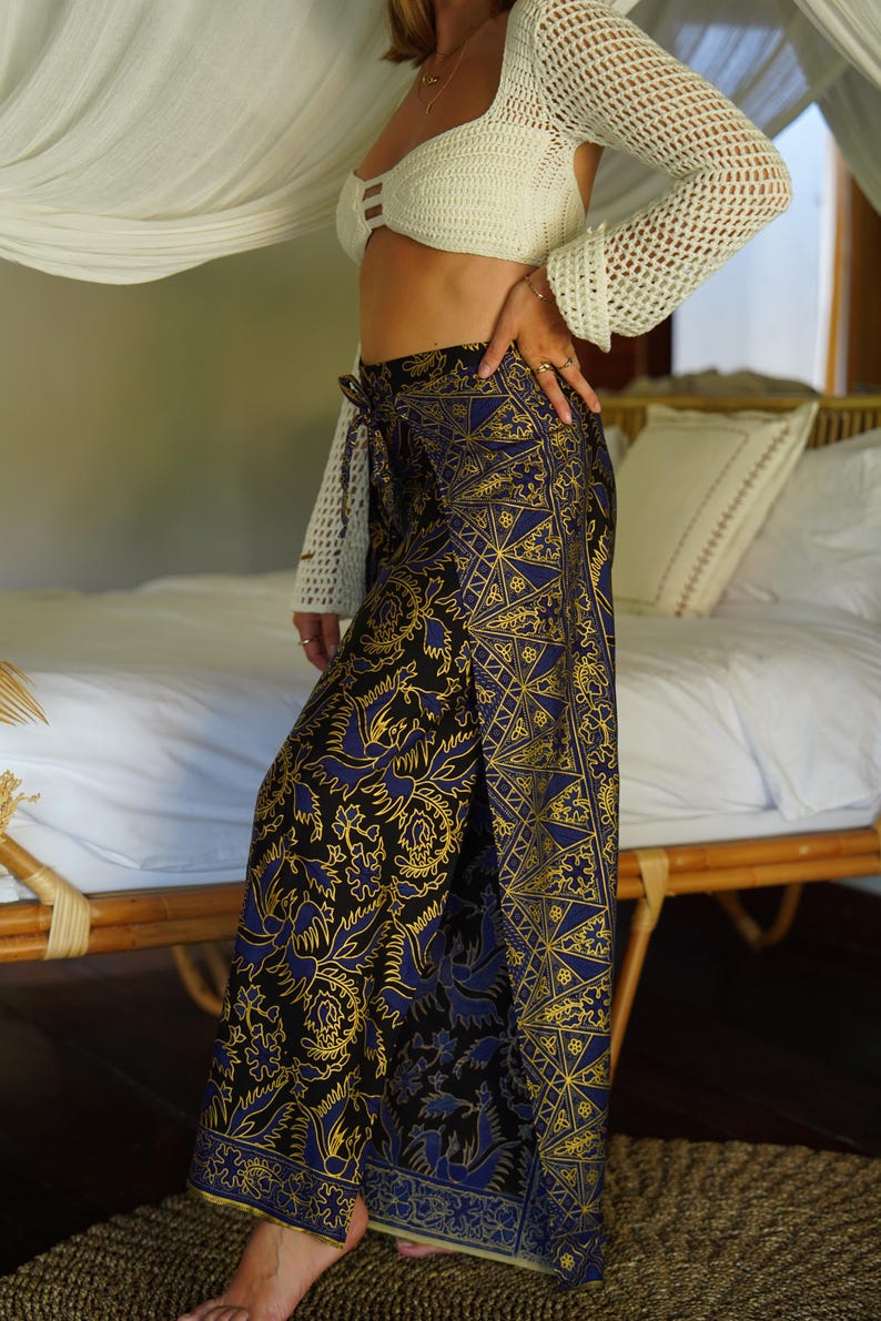 May include: A woman wearing a white crochet top and a long, blue and gold patterned wrap skirt. The skirt has a floral design and is tied at the waist.