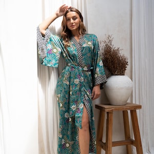 Op de afbeelding: Een turquoise kimono met bloemenprint, wijde mouwen en bijpassende riem. De kimono heeft een contrasterend zwart-wit patroon op de manchetten en kraag. De lange kimono is knielang en heeft een hoge split.