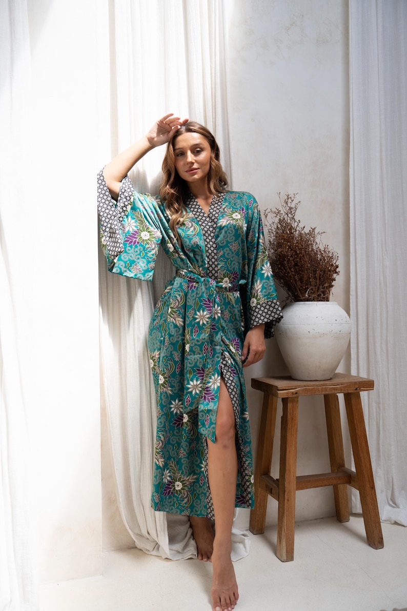 Op de afbeelding: Een turquoise kimono met bloemenprint, wijde mouwen en bijpassende riem. De kimono heeft een contrasterend zwart-wit patroon op de manchetten en kraag. De lange kimono is knielang en heeft een hoge split.