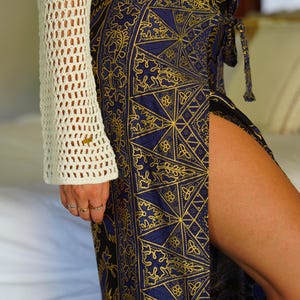 May include: A woman wearing a white crochet top and a blue and gold patterned skirt with a side slit. The skirt has a geometric design with intricate details.