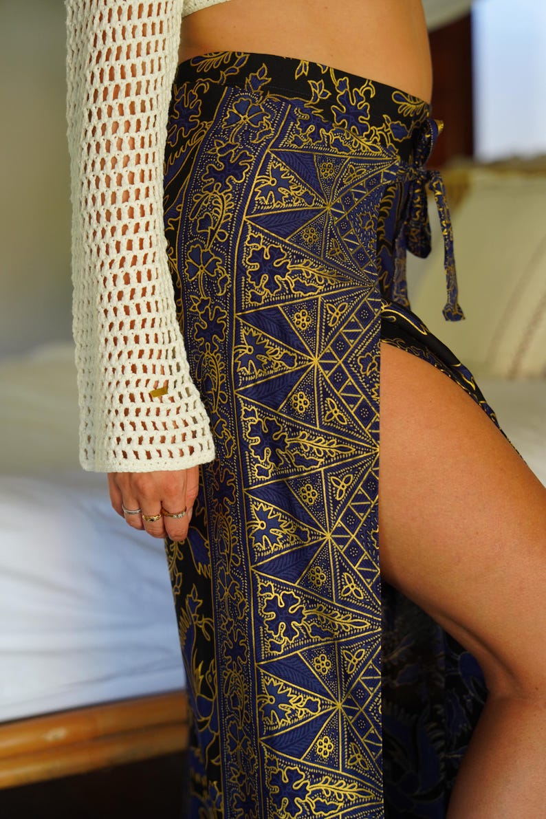 May include: A woman wearing a white crochet top and a blue and gold patterned skirt with a side slit. The skirt has a geometric design with intricate details.