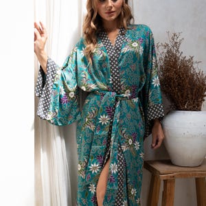 Op de afbeelding: Een turquoise kimono-stijl badjas met een bloemenpatroon in wit, paars en groen. De badjas heeft wijde mouwen met een contrasterende zwart-witte geometrische print. Het wordt in de taille vastgebonden met een bijpassende riem.