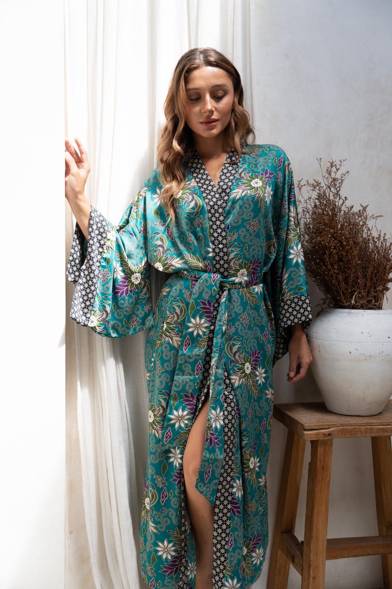 Op de afbeelding: Een turquoise kimono-stijl badjas met een bloemenpatroon in wit, paars en groen. De badjas heeft wijde mouwen met een contrasterende zwart-witte geometrische print. Het wordt in de taille vastgebonden met een bijpassende riem.