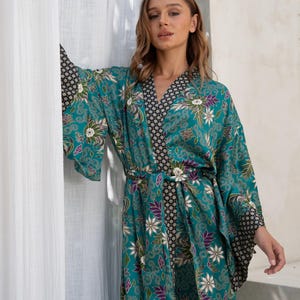 Op de afbeelding: Een turquoise kimono-achtige badjas met een bloemenpatroon in wit, paars en groen. De badjas heeft een zwart-wit geometrisch patroon op de kraag en manchetten. Het is in de taille vastgebonden met een bijpassende riem.
