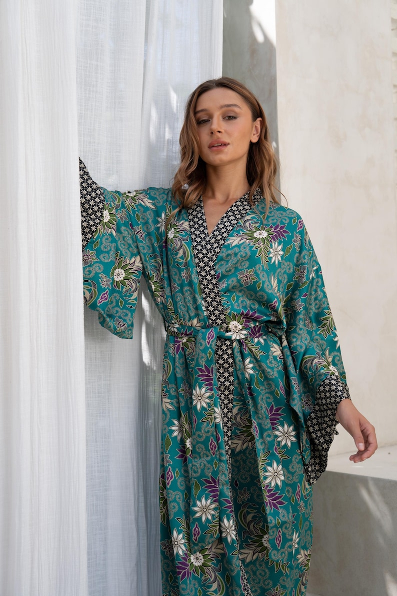 Op de afbeelding: Een turquoise kimono-achtige badjas met een bloemenpatroon in wit, paars en groen. De badjas heeft een zwart-wit geometrisch patroon op de kraag en manchetten. Het is in de taille vastgebonden met een bijpassende riem.
