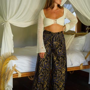 May include: A woman wearing a white crochet top with a black and gold patterned pants. The pants have a wide leg and a high waist. The woman is standing in front of a bed with white linens.