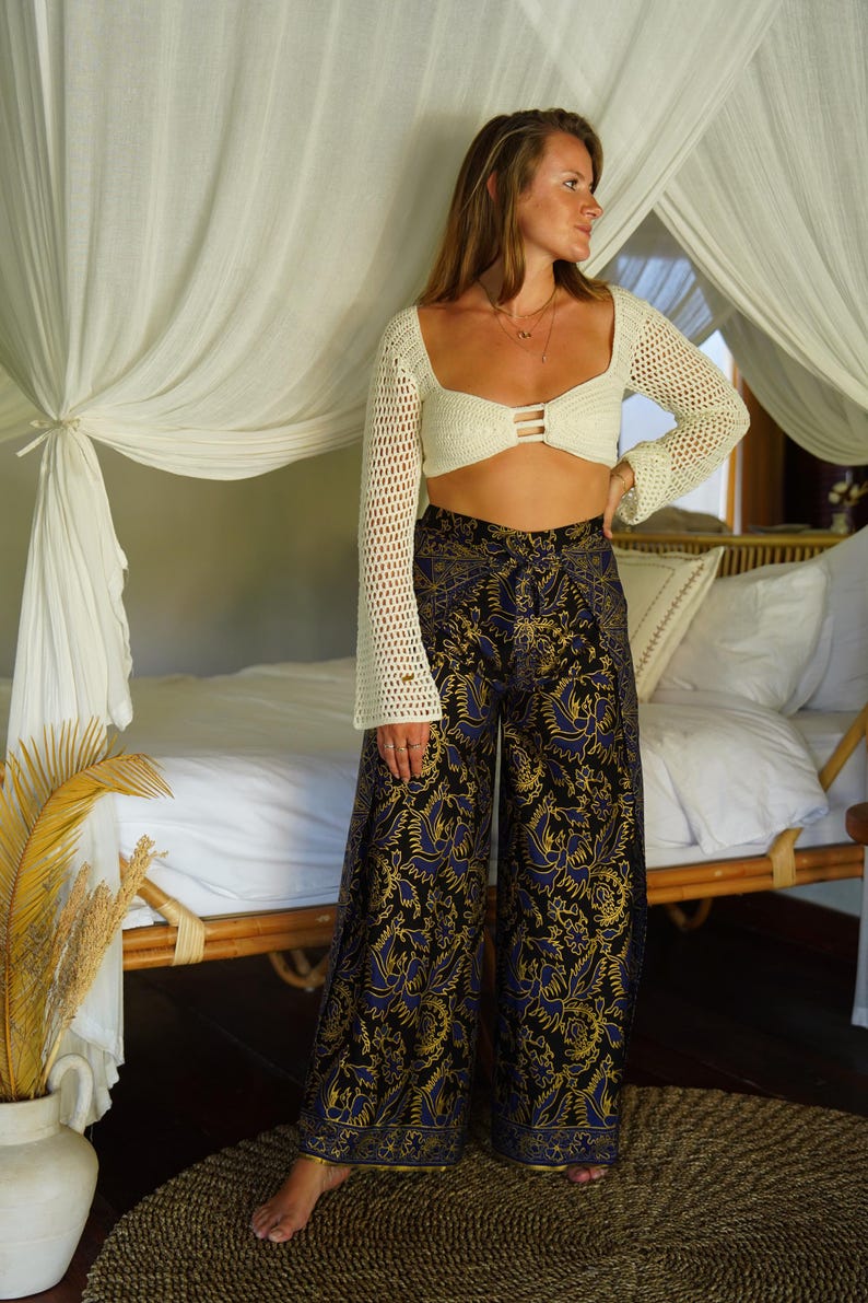 May include: A woman wearing a white crochet top with a black and gold patterned pants. The pants have a wide leg and a high waist. The woman is standing in front of a bed with white linens.