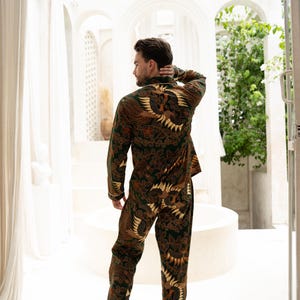 Puede incluir: Un conjunto de pijama de dos piezas con un estampado batik marr&oacute;n y verde oscuro con un dise&ntilde;o de p&aacute;jaro. La parte superior de manga larga y los pantalones tienen ribetes blancos. La persona est&aacute; de pie en una habitaci&oacute;n blanca y luminosa.