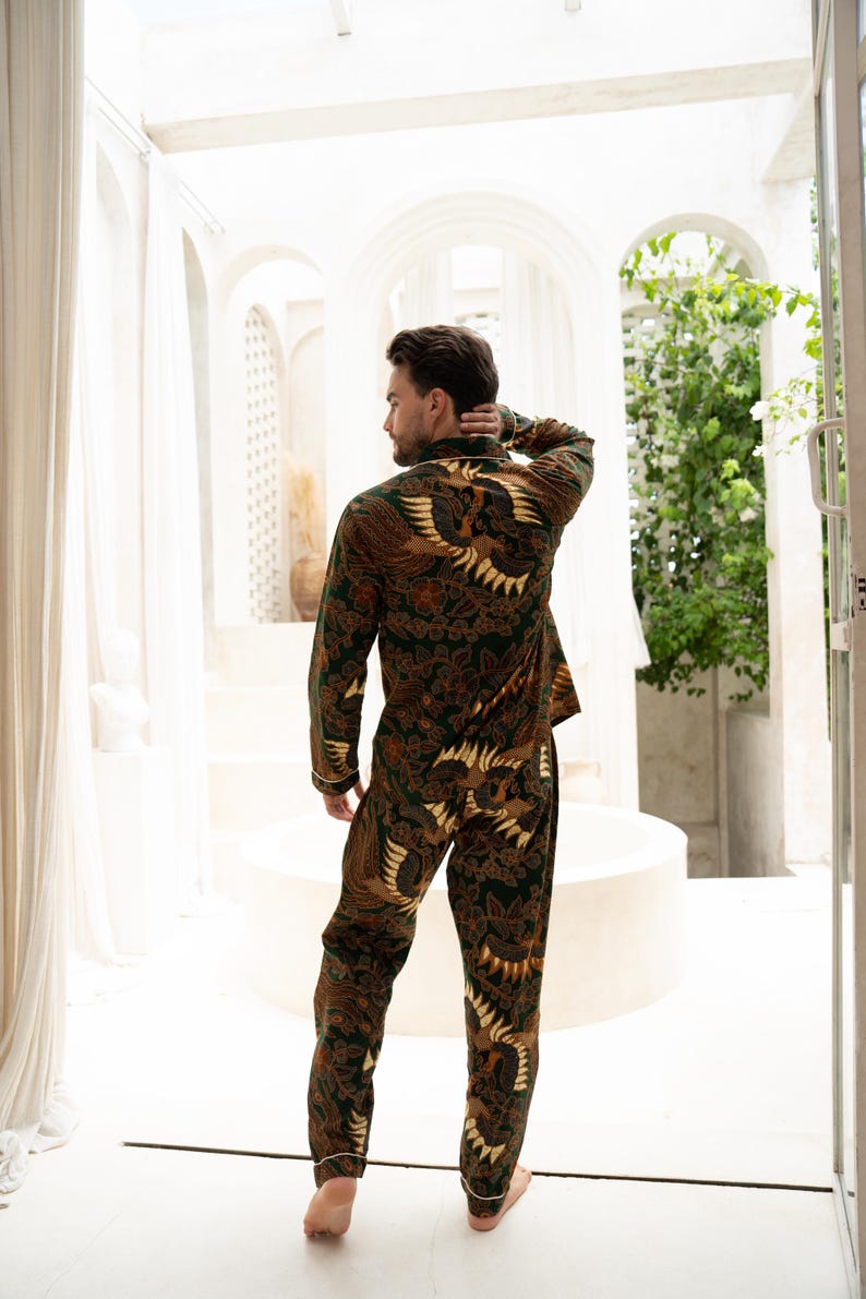 Puede incluir: Un conjunto de pijama de dos piezas con un estampado batik marr&oacute;n y verde oscuro con un dise&ntilde;o de p&aacute;jaro. La parte superior de manga larga y los pantalones tienen ribetes blancos. La persona est&aacute; de pie en una habitaci&oacute;n blanca y luminosa.