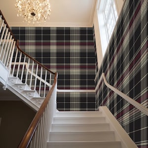 May include: A staircase with white steps and a wooden handrail ascends towards a wall covered in a black, burgundy, and cream plaid pattern. A chandelier hangs from the ceiling, illuminating the space.