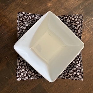 May include: A white square ceramic bowl resting inside a dark brown fabric bowl cozy. The fabric features a repeating pattern of small white flowers and thin stems. The bowl is positioned at an angle, showing its smooth interior and edges.