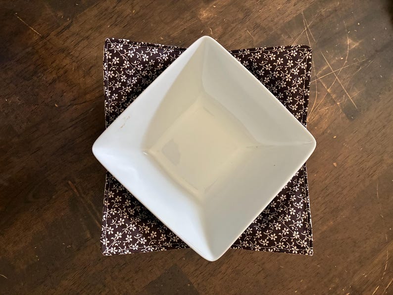 May include: A white square ceramic bowl resting inside a dark brown fabric bowl cozy. The fabric features a repeating pattern of small white flowers and thin stems. The bowl is positioned at an angle, showing its smooth interior and edges.