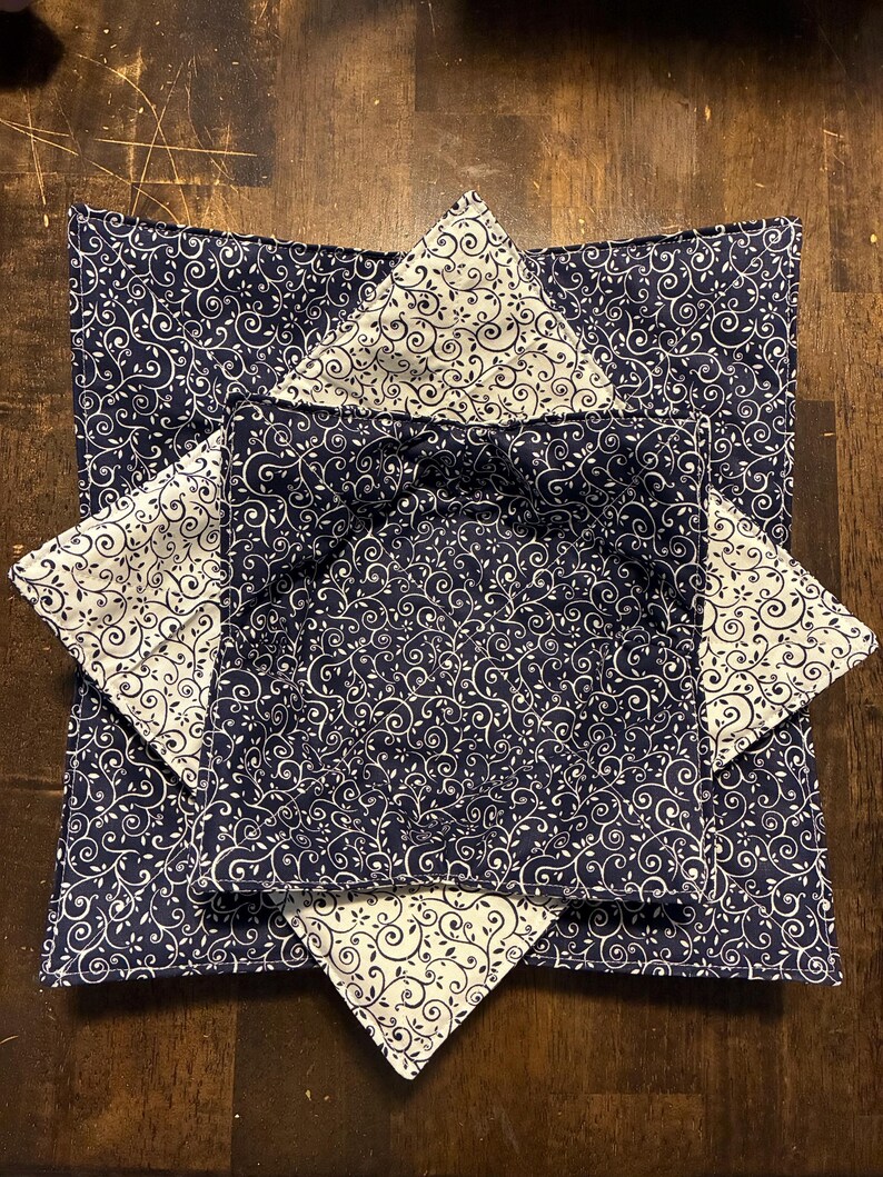 May include: A square fabric bowl cozy featuring a navy blue background with a white swirling vine pattern on the exterior and a white background with a navy blue swirling vine pattern on the interior, constructed with quilted stitching.