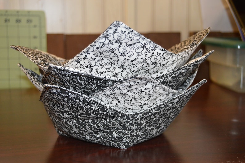 May include: A fabric bowl cozy featuring a black and white floral scroll pattern. The square-shaped holder has four pointed corners and is constructed with quilted fabric, displaying a consistent intricate vine design on both the interior and exterior.