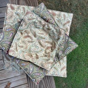 May include: A set of three square fabric bowl cozies featuring a repeating paisley pattern in shades of tan, brown, green, blue, and red. The quilted fabric items are stacked on top of each other, displaying a consistent print across all surfaces.