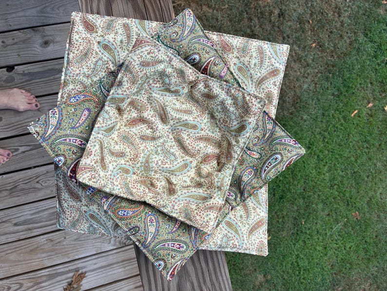 May include: A set of three square fabric bowl cozies featuring a repeating paisley pattern in shades of tan, brown, green, blue, and red. The quilted fabric items are stacked on top of each other, displaying a consistent print across all surfaces.