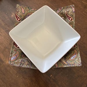 May include: A white square ceramic bowl resting inside a fabric bowl cozy. The cozy features a multi-colored paisley pattern with shades of olive green, burgundy, and blue on a textured fabric base, providing a snug fit for the bowl.