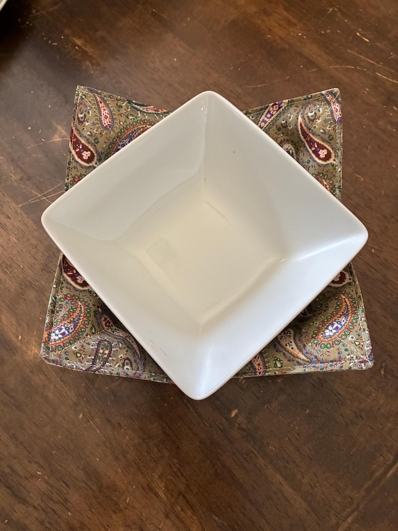 May include: A white square ceramic bowl resting inside a fabric bowl cozy. The cozy features a multi-colored paisley pattern with shades of olive green, burgundy, and blue on a textured fabric base, providing a snug fit for the bowl.