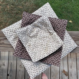 May include: A stack of three square fabric bowl cozies featuring a repeating small floral pattern. The top and bottom cozies are cream with brown flowers, while the middle cozy is brown with cream flowers, all with quilted stitching.