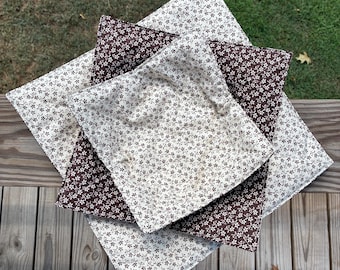 May include: A stack of three square fabric bowl cozies featuring a repeating small floral pattern. The top and bottom cozies are cream with brown flowers, while the middle cozy is brown with cream flowers, all with quilted stitching.