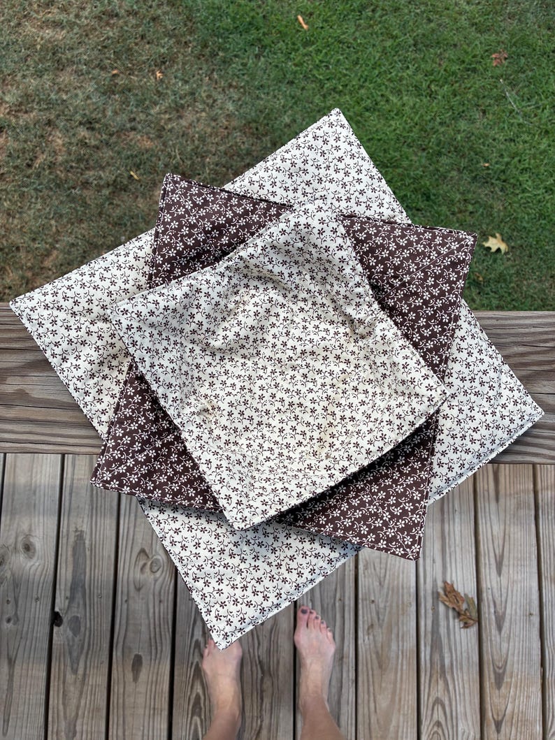 May include: A stack of three square fabric bowl cozies featuring a repeating small floral pattern. The top and bottom cozies are cream with brown flowers, while the middle cozy is brown with cream flowers, all with quilted stitching.