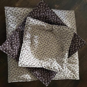 May include: Two square fabric bowl cozies stacked on top of each other. The top cozy features a cream background with a dark brown floral pattern, while the bottom cozy features a dark brown background with a cream floral pattern. Both have quilted stitching.