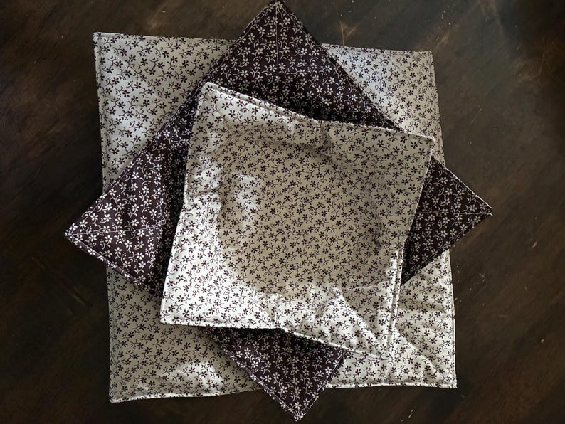 May include: Two square fabric bowl cozies stacked on top of each other. The top cozy features a cream background with a dark brown floral pattern, while the bottom cozy features a dark brown background with a cream floral pattern. Both have quilted stitching.
