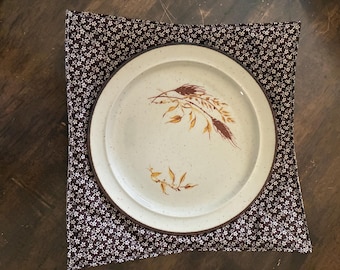 May include: A round, off-white ceramic plate with a speckled finish and a brown wheat stalk illustration, centered inside a square, quilted fabric bowl cozy featuring a dark brown background with a small, repeating white floral pattern.