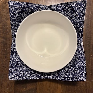 May include: A square fabric bowl cozy featuring a dark navy blue background with a repeating white swirling vine and leaf pattern. The cozy has four folded corners and holds a plain white ceramic bowl centered within its quilted fabric structure.