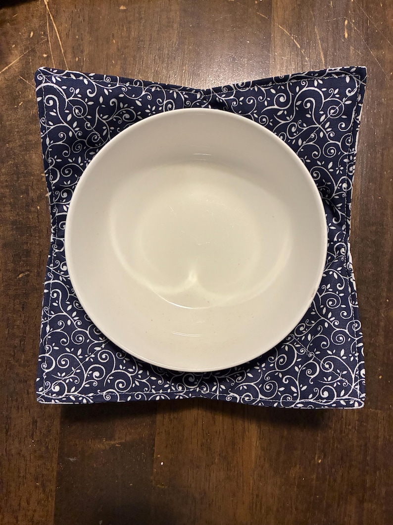 May include: A square fabric bowl cozy featuring a dark navy blue background with a repeating white swirling vine and leaf pattern. The cozy has four folded corners and holds a plain white ceramic bowl centered within its quilted fabric structure.