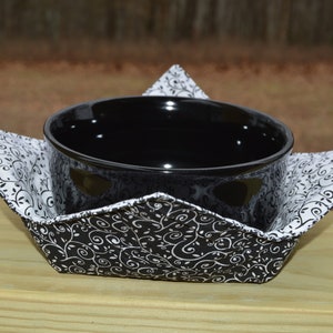 May include: A black ceramic bowl sits inside a fabric bowl cozy. The cozy features a black and white swirling vine pattern on the exterior and a white fabric with a black swirling vine pattern on the interior, with four pointed corners.