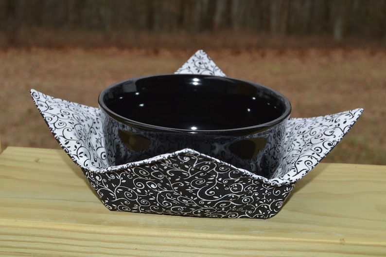 May include: A black ceramic bowl sits inside a fabric bowl cozy. The cozy features a black and white swirling vine pattern on the exterior and a white fabric with a black swirling vine pattern on the interior, with four pointed corners.